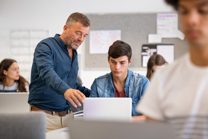 Professor assisting college student with laptop in classroom during computer lesson. Teacher talking and explaining to guy. Mature man lecturer helping high school teen with laptop during lecture.